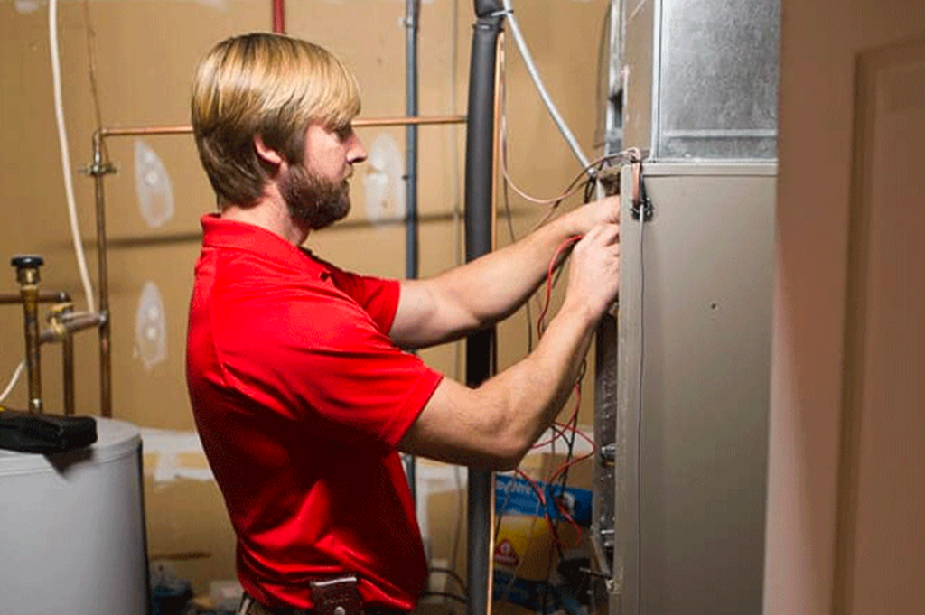 A man in a red shirt with a beard and blond hair is working on an electrical panel or appliance. He's focused on connecting or checking wires inside the unit, positioned in a room with beige walls and other equipment around.